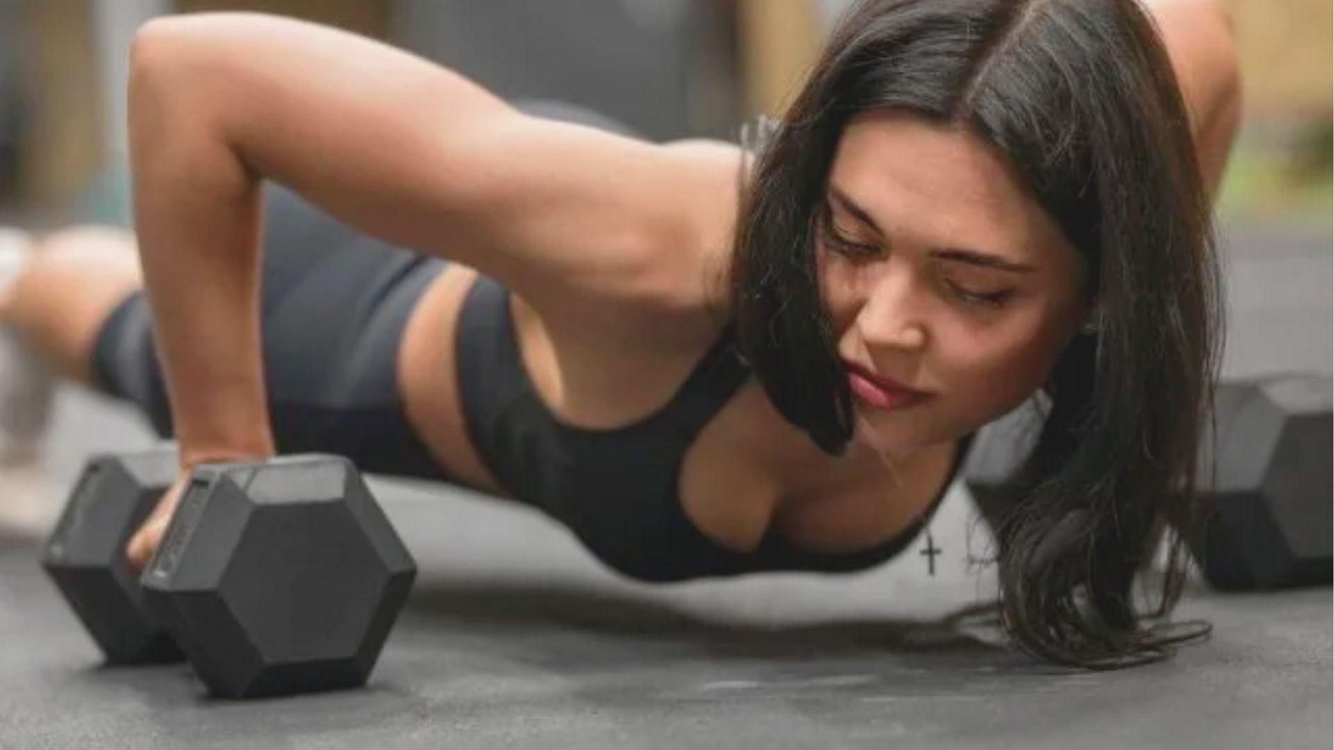 Woman in a black sports bra performing a deep push-up while gripping two dumbbells on the gym floor.