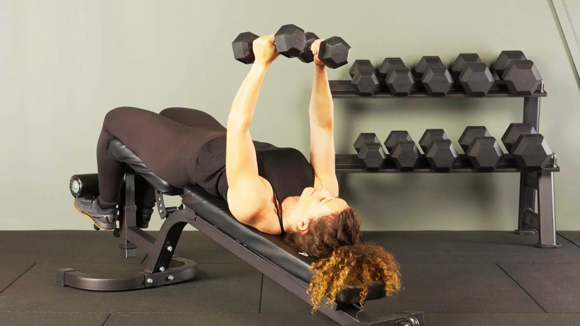 A woman holds dumbbells at the top of a decline bench press, with feet locked under pads.