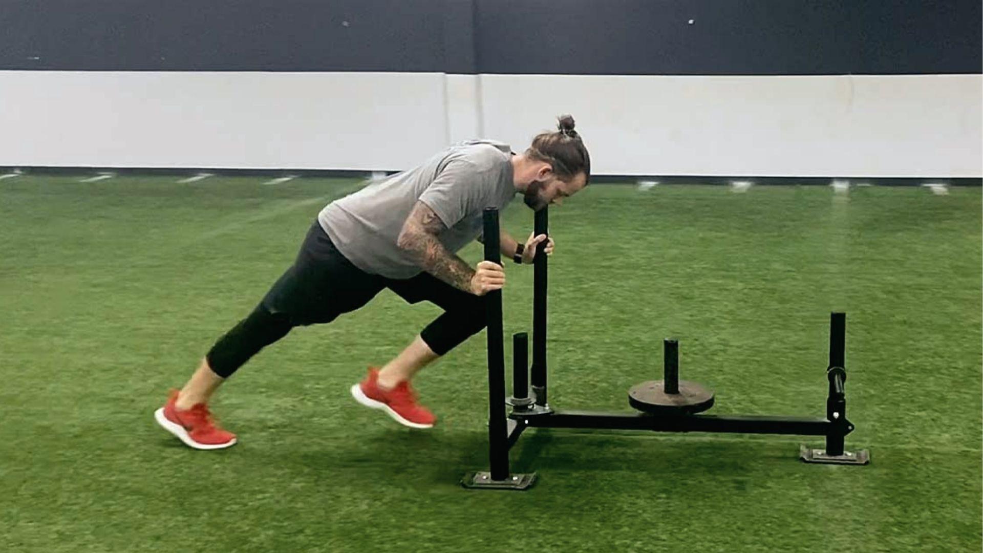 A man in gray tee and red shoes pushes sleds on an indoor turf field, body angled low for max force in sled workout.