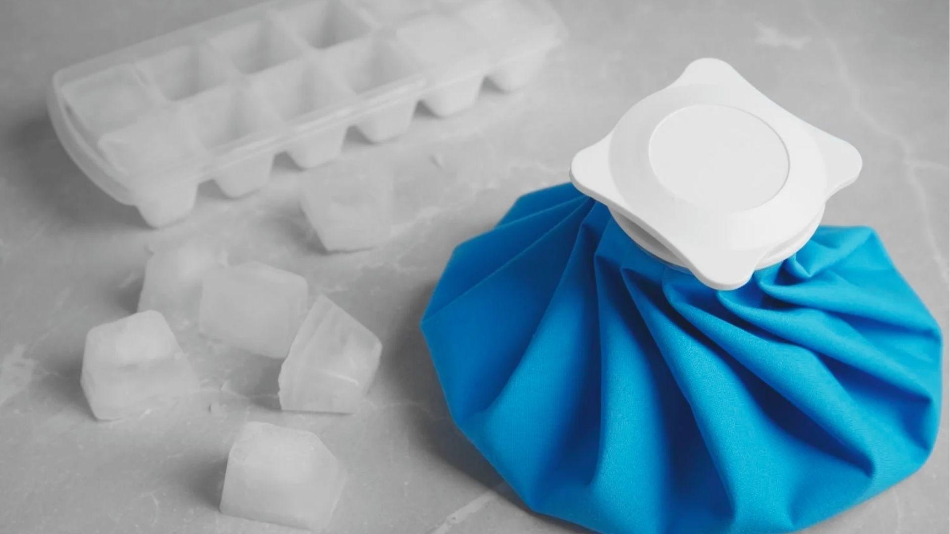 Blue reusable ice pack next to scattered ice cubes and a plastic ice tray on a marble counter.