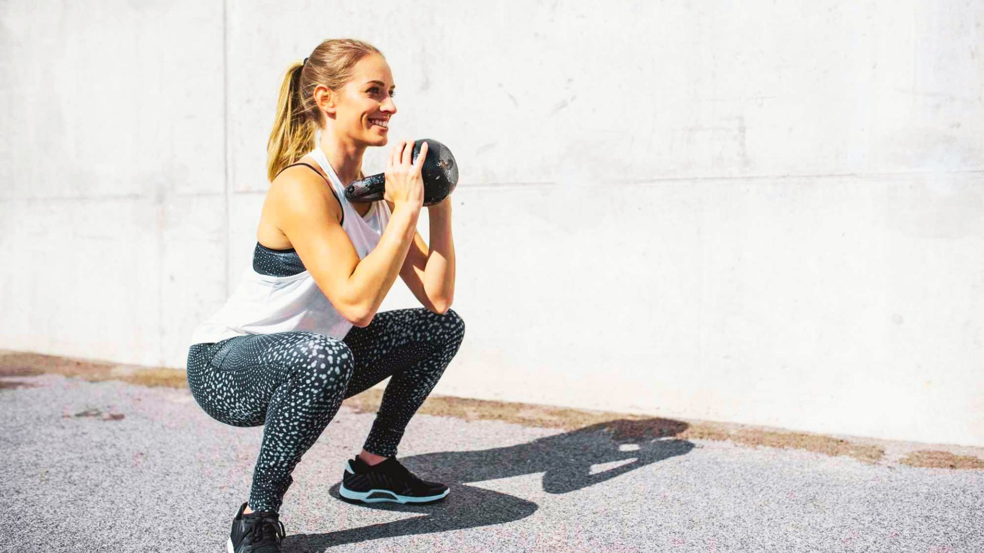 Smiling woman doing a kettlebell goblet squat outdoors against a white wall, holding the weight close to her chest.