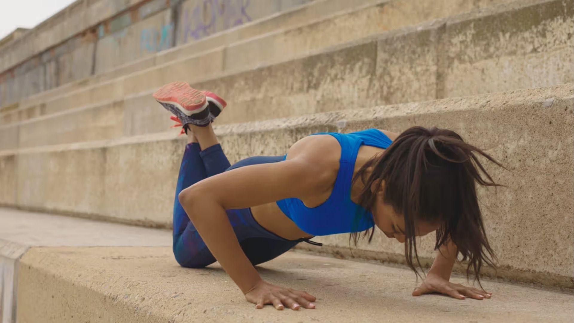 Woman doing a modified knee push-up on a concrete ledge outdoors in a blue sports bra.