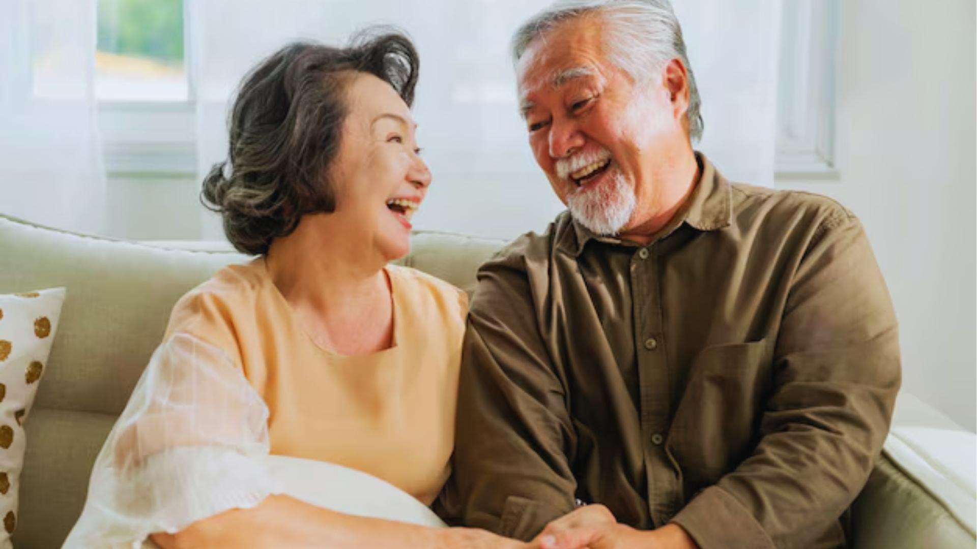 Elderly Asian couple sits on beige couch laughing together, holding hands in bright living room for heart health connection.