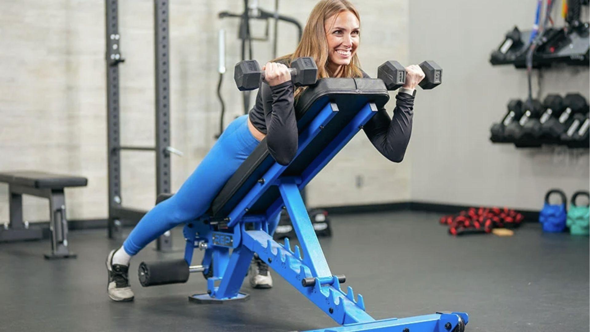 Muscular woman does dumbbell spider curls lying on incline bench, elbows forward, biceps fully engaged.
