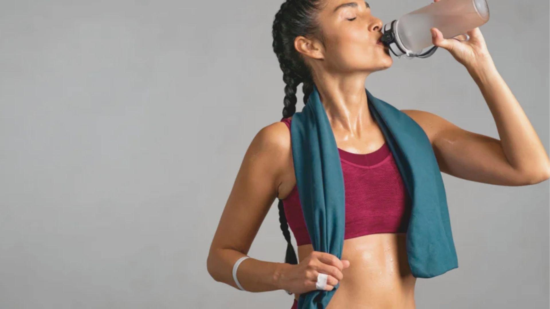 Athletic woman in maroon sports bra drinks from a bottle with a towel around her neck, appearing sweaty after exercise.