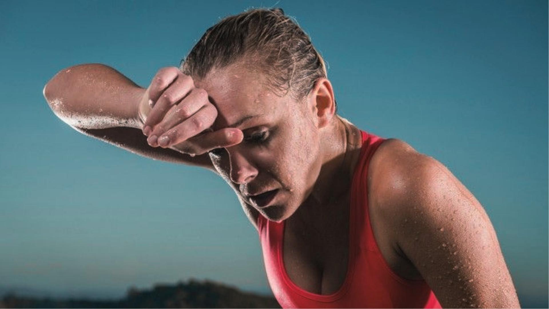 Woman in red tank top leans forward outdoors, wiping sweat from forehead with forearm after intense cardio.