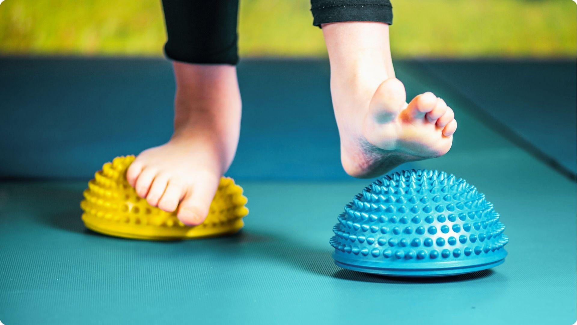 Bare feet balance on yellow and blue spiky dome balance pods placed on a teal exercise mat.