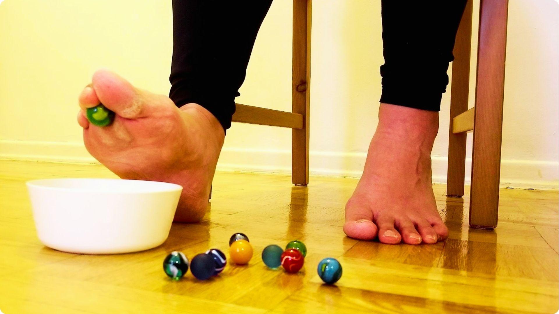 Close-up of bare feet doing a marble pickup, with toes gripping a green marble into a white bowl.