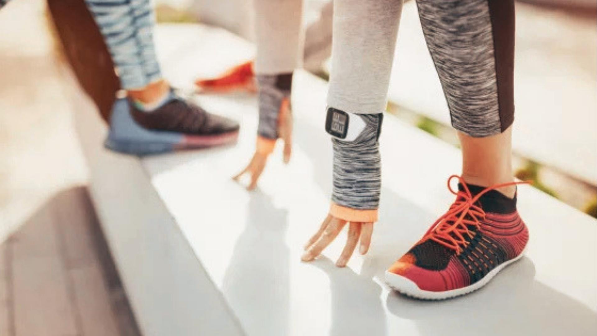 Three people in workout leggings crouch in starting position on track, wearing running shoes and fitness tracker.