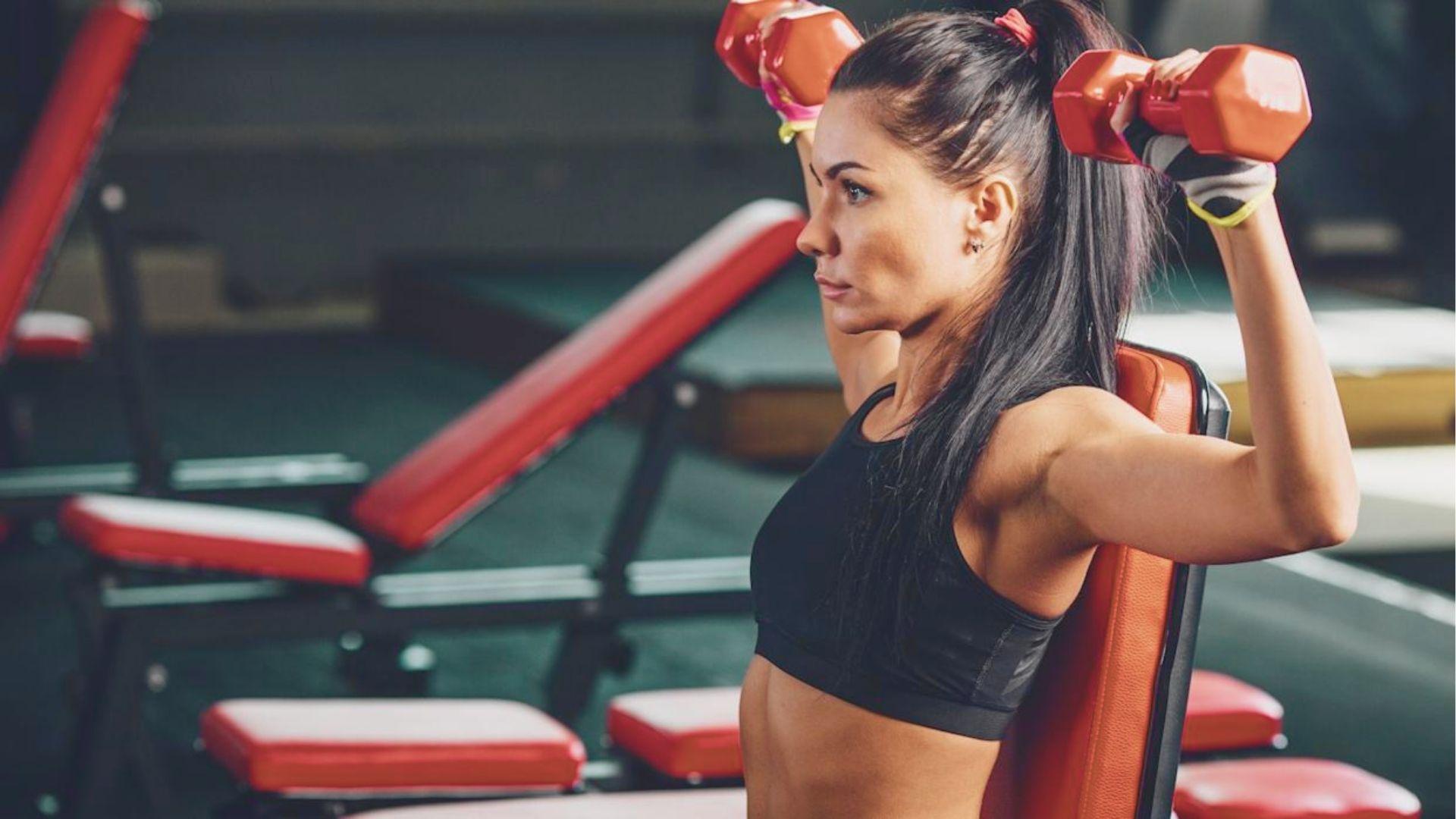 Woman with a high ponytail sitting on a bench, pressing red dumbbells overhead with elbows bent.