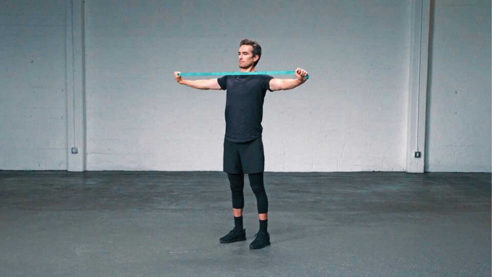 Man in black athletic wear performs resistance band pull-apart at chest height to strengthen rotator cuff muscles.