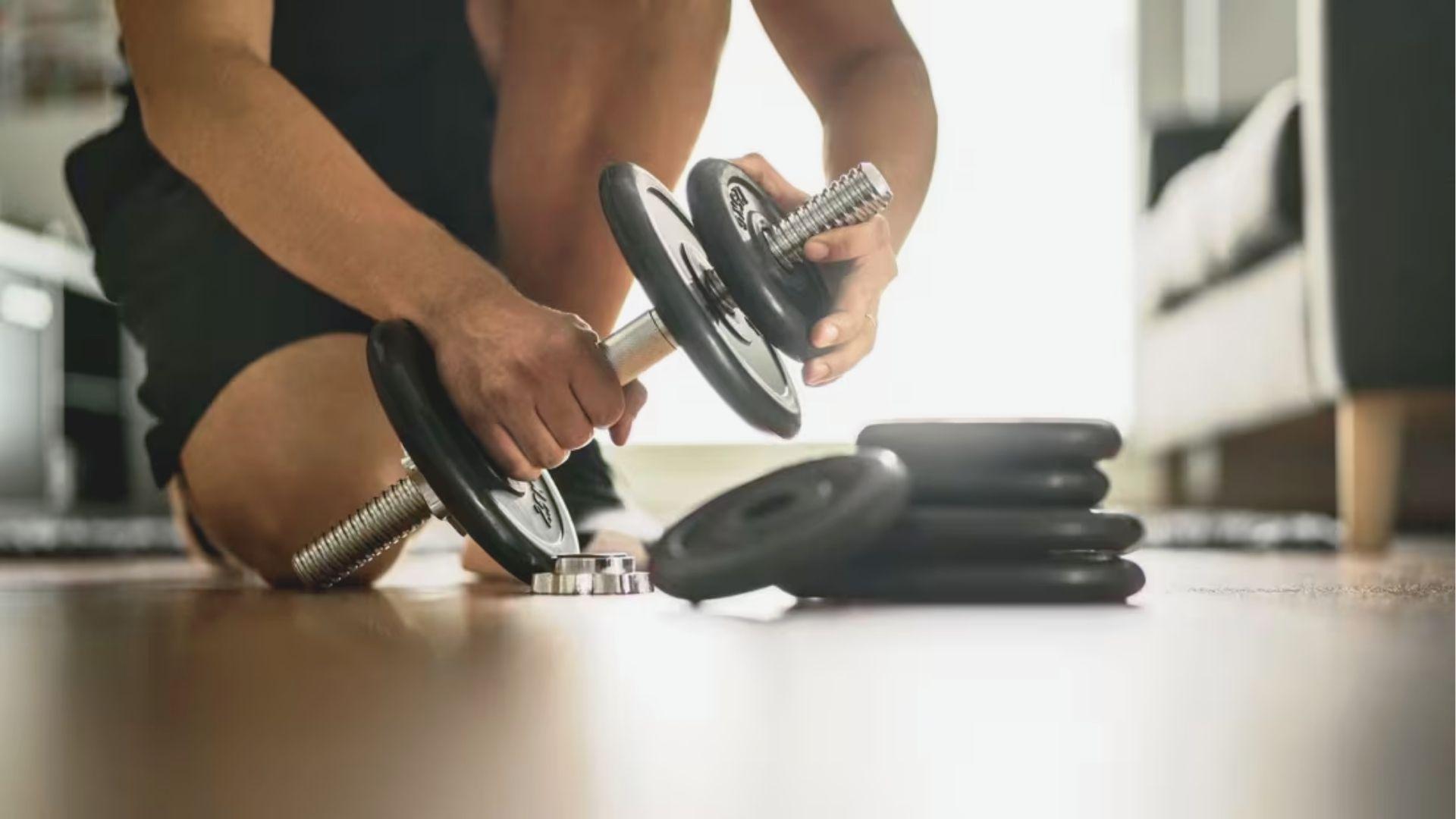 Person kneeling on a gym floor adjusts weight plates on an adjustable dumbbell for their next set.