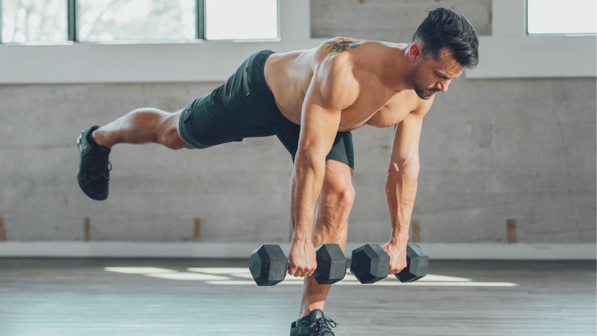 Shirtless man performs a single-leg dumbbell Romanian deadlift in a gym, holding two dumbbells.