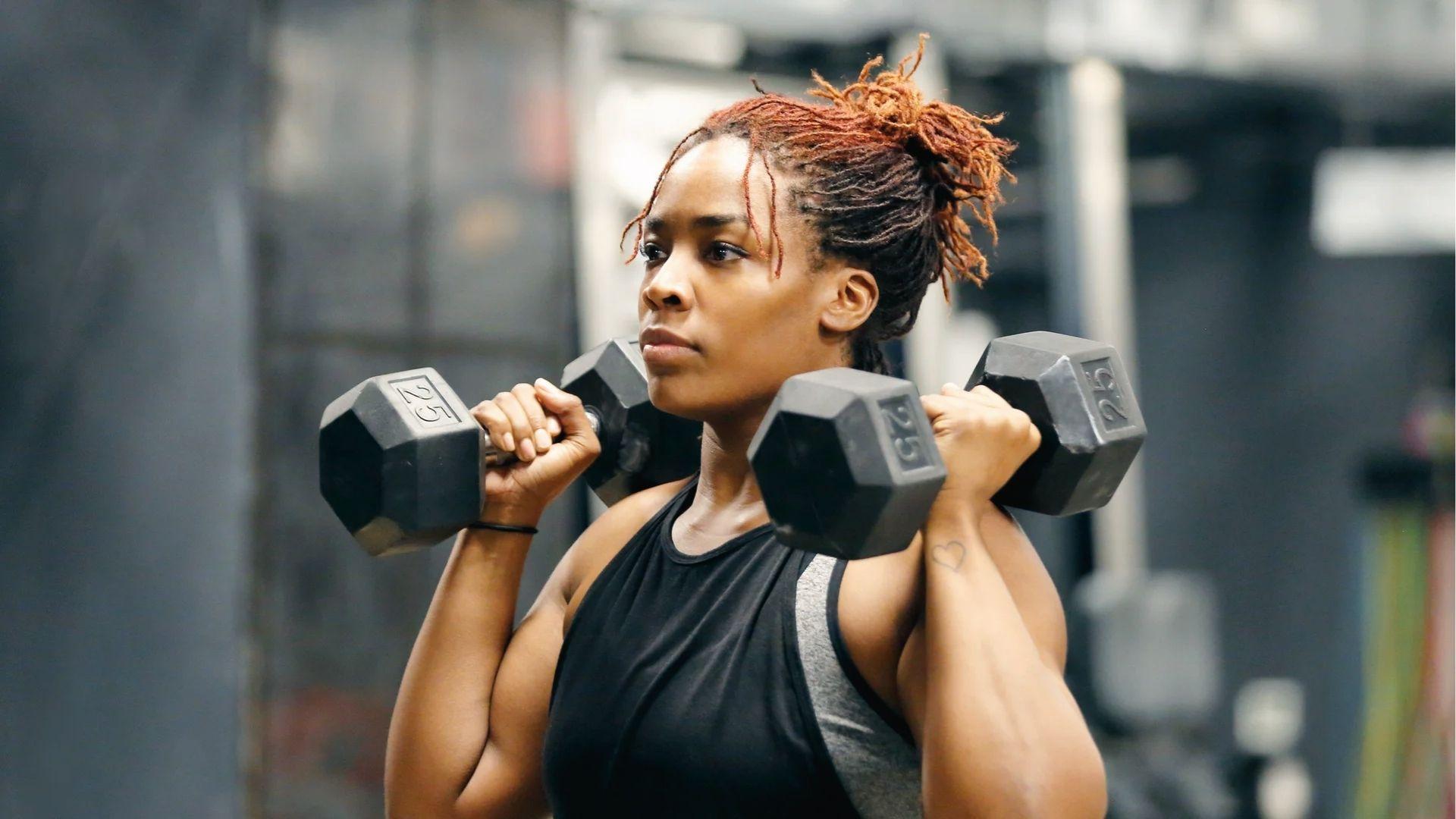 A woman holding 25lb dumbbells at shoulders in the gym, showing progressive overload builds muscular strength.