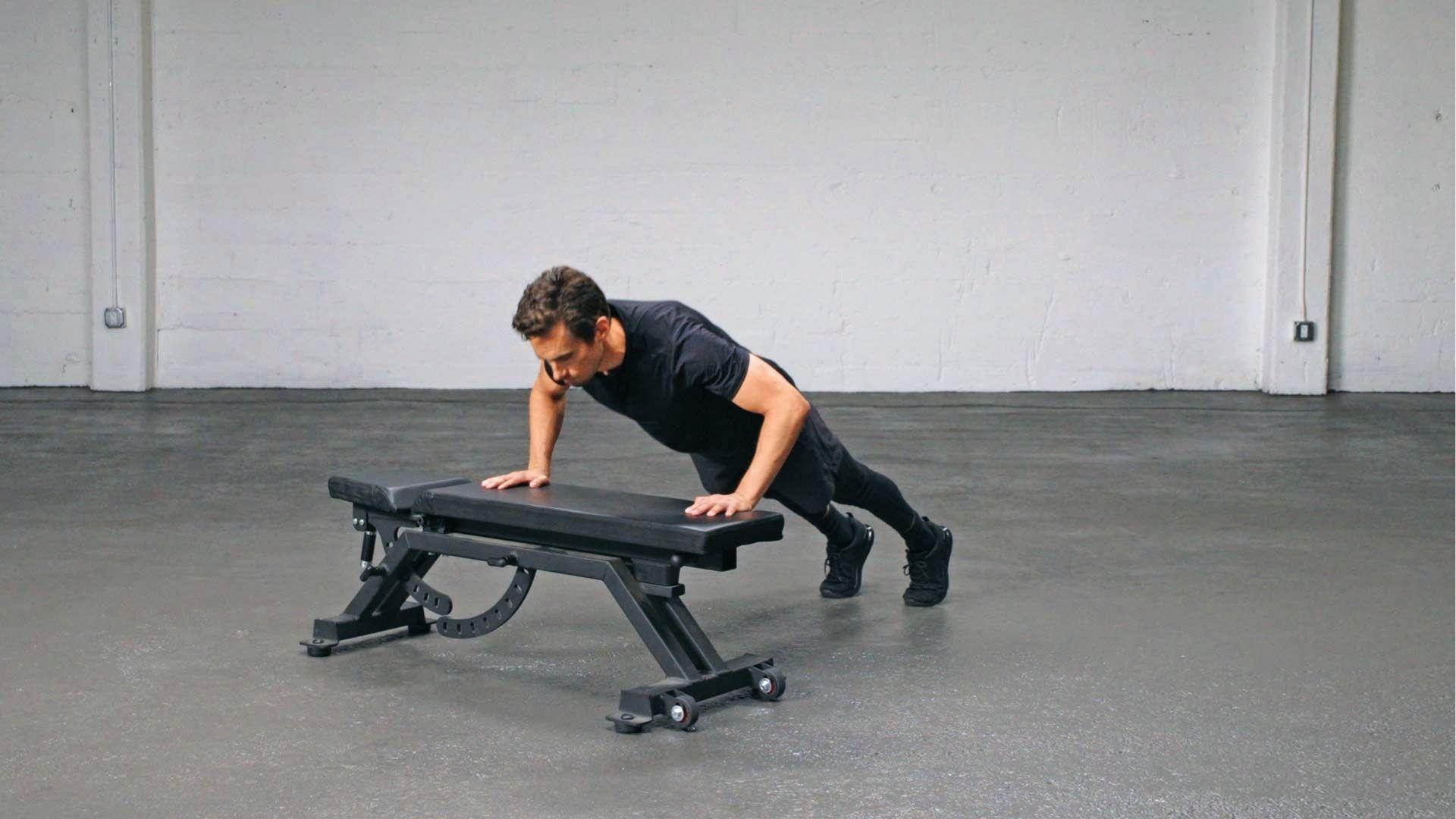 Man in black outfit holds incline push up position on flat bench with gray floor and white walls