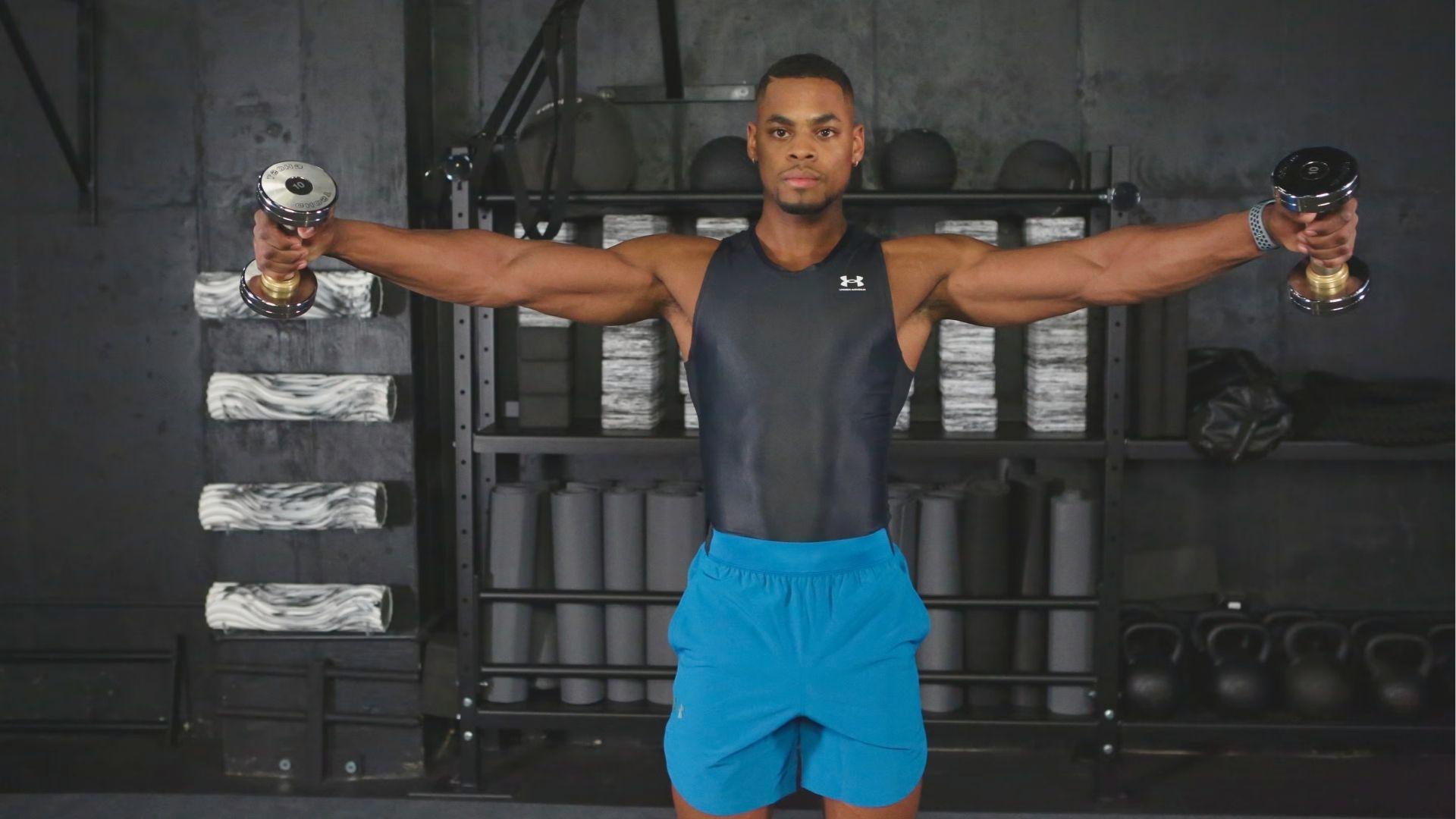 A man in a black tank top and blue shorts holds dumbbells at shoulder level during a lateral raise.