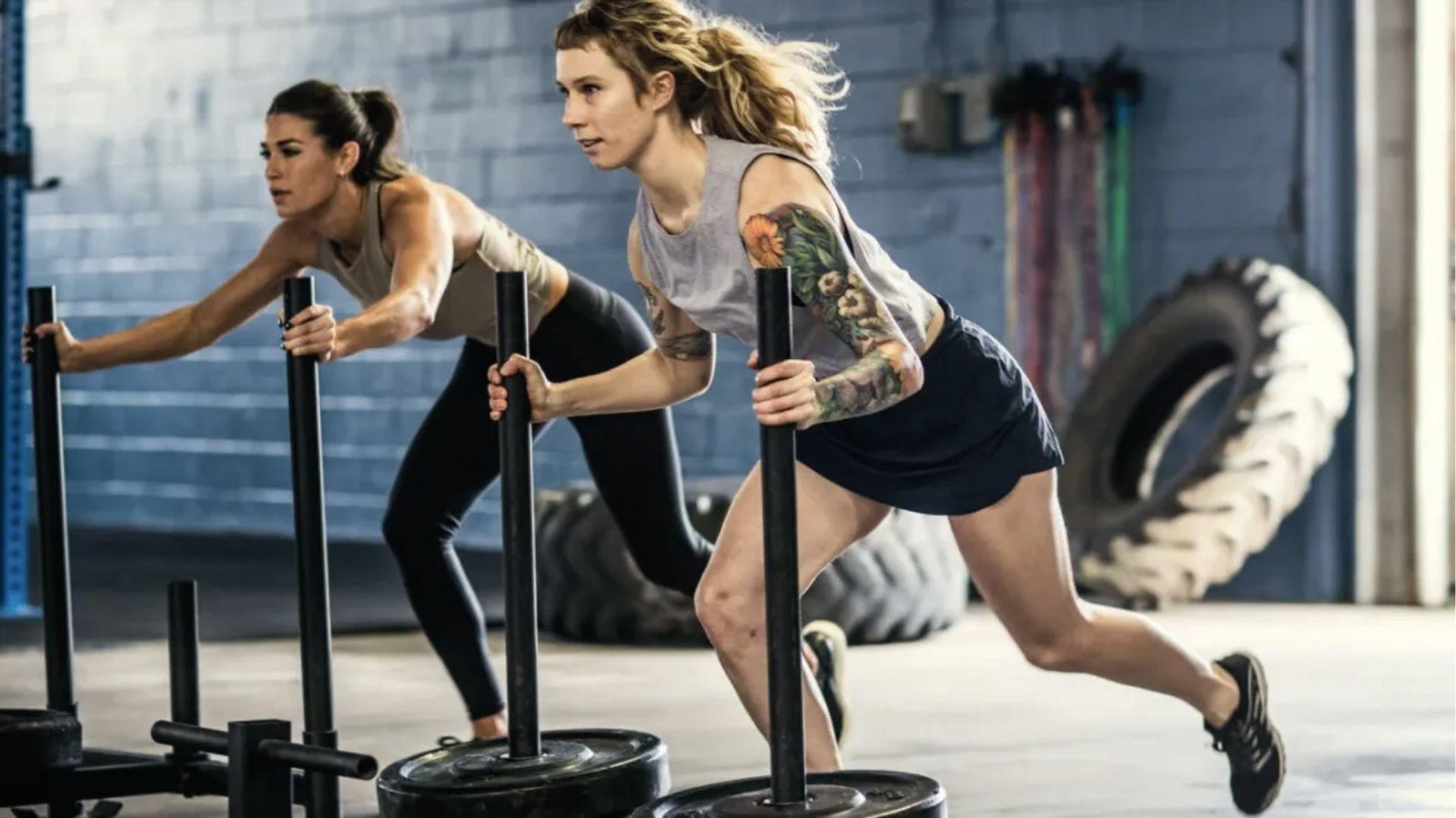 Two women push weighted sleds side by side in the gym, showcasing partner training and full-body sled push effort.