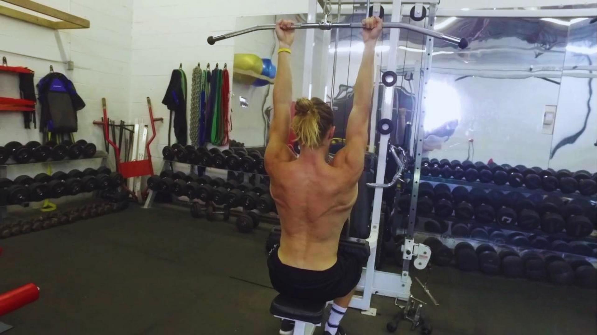 Shirtless man with blonde bun starts lat pulldown with overhand grip showing engaged back muscles in dumbbell-filled gym.