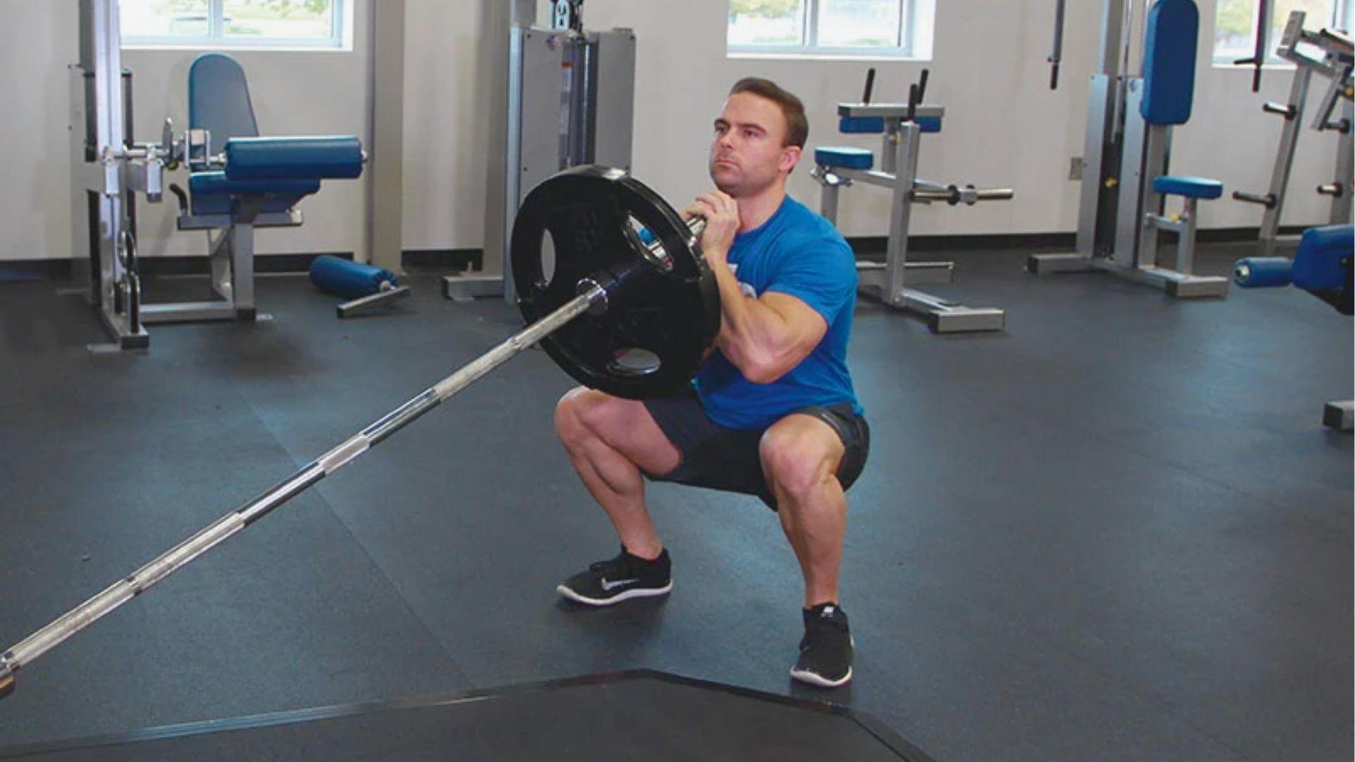 A man in a blue shirt and black shorts holds the landmine barbell at chest level in a deep squat position.