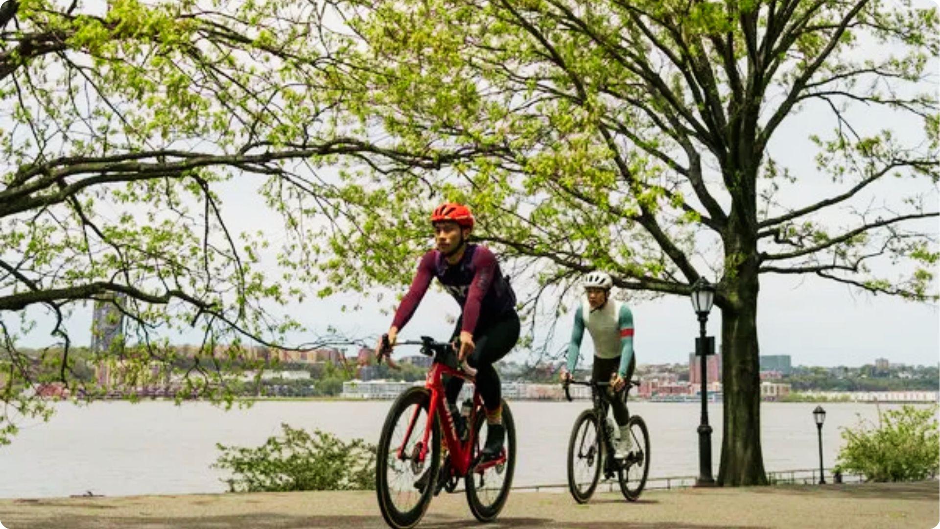 Two cyclists ride road bikes along a waterfront path with trees and a city skyline.