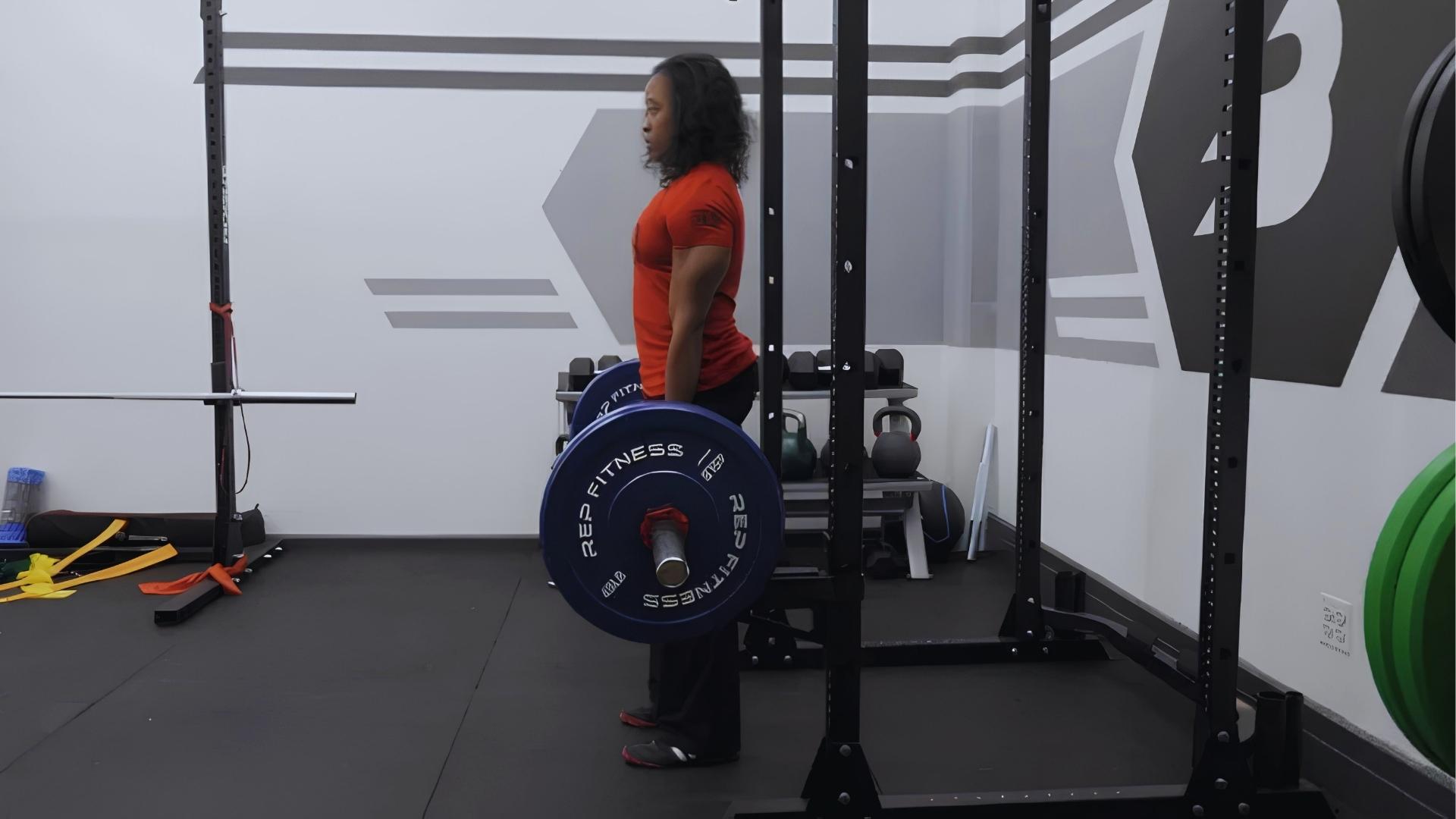 Woman in red shirt performs barbell Romanian deadlift inside squat rack with blue plates in modern home gym setup.