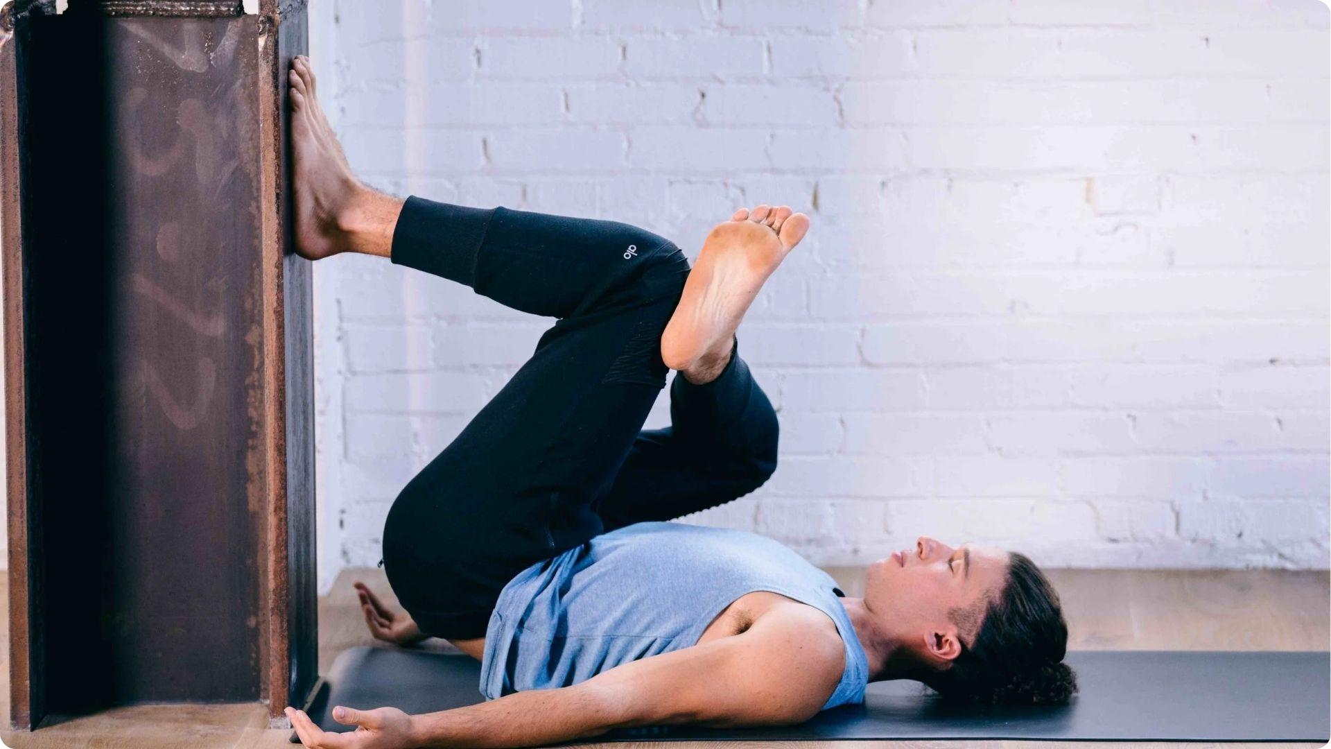 Man lying on his back on a mat with one foot pressed against a wall and the opposite ankle crossed over the thigh.