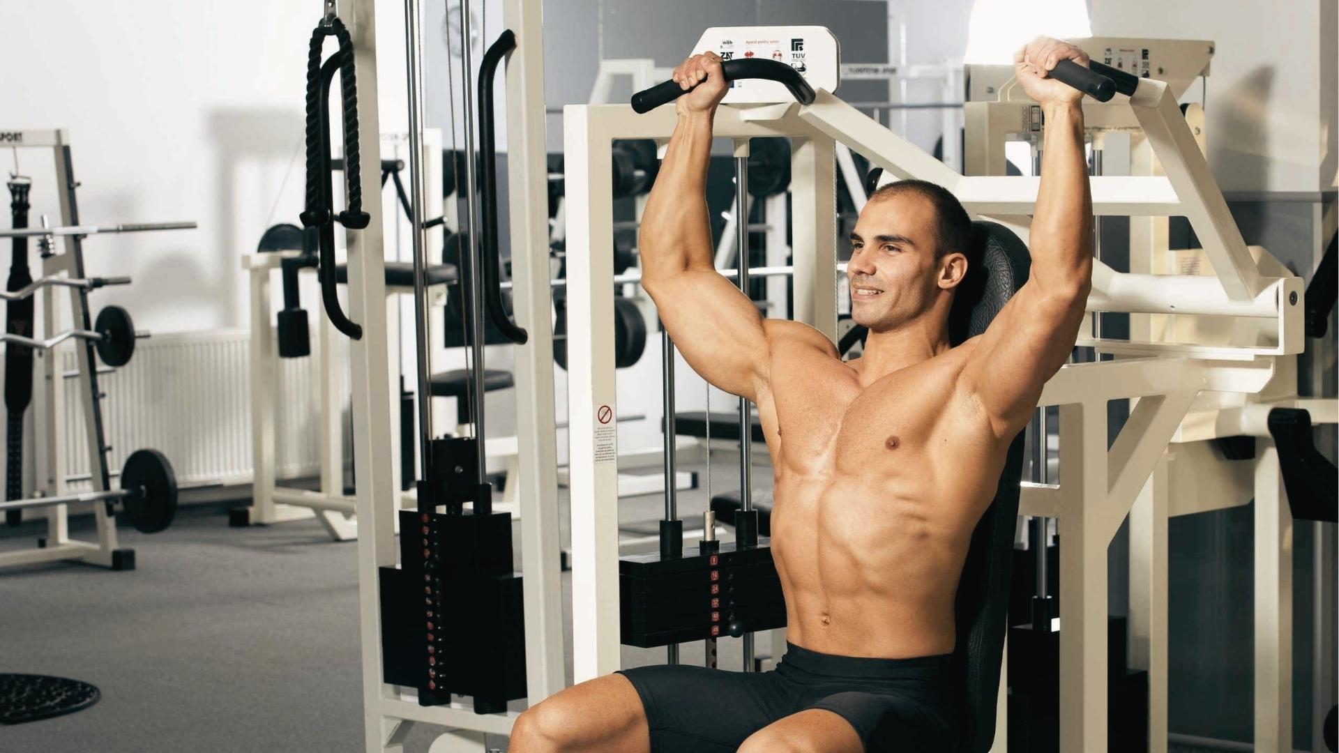 Shirtless man with defined abs uses a seated shoulder press machine in a bright gym, arms fully extended overhead.