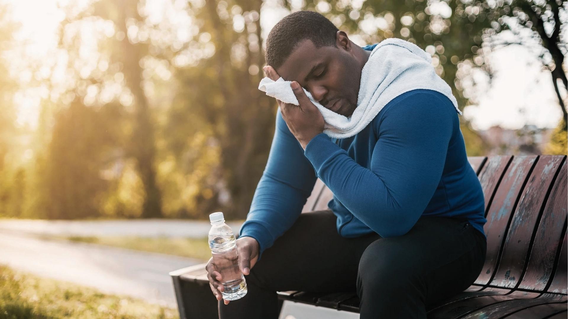 Exhausted man in blue t-shirt sits on park bench wiping sweat with towel, holding water bottle after workout.