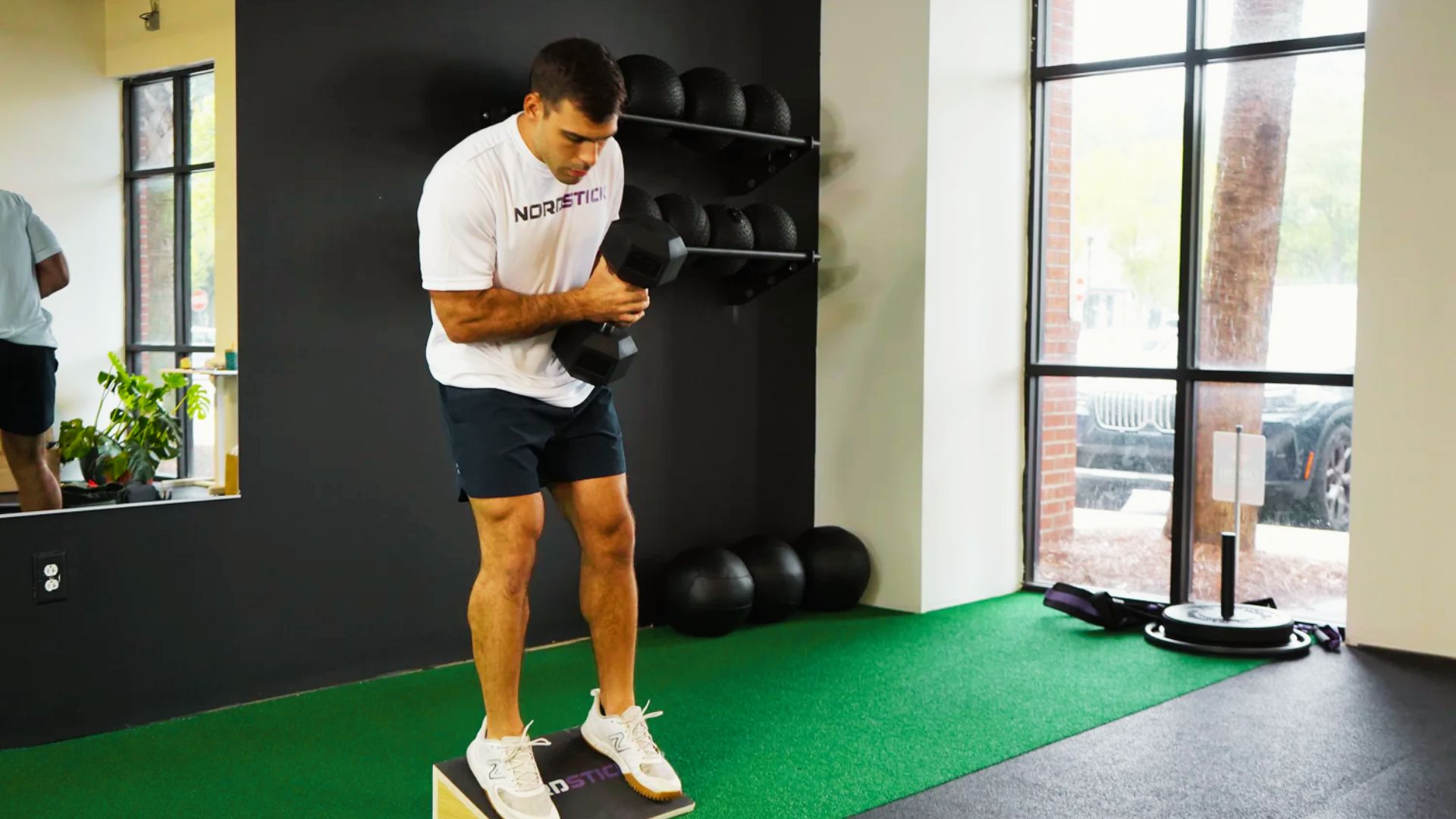 A man stands with both feet on a slant board while holding a dumbbell close to his chest.