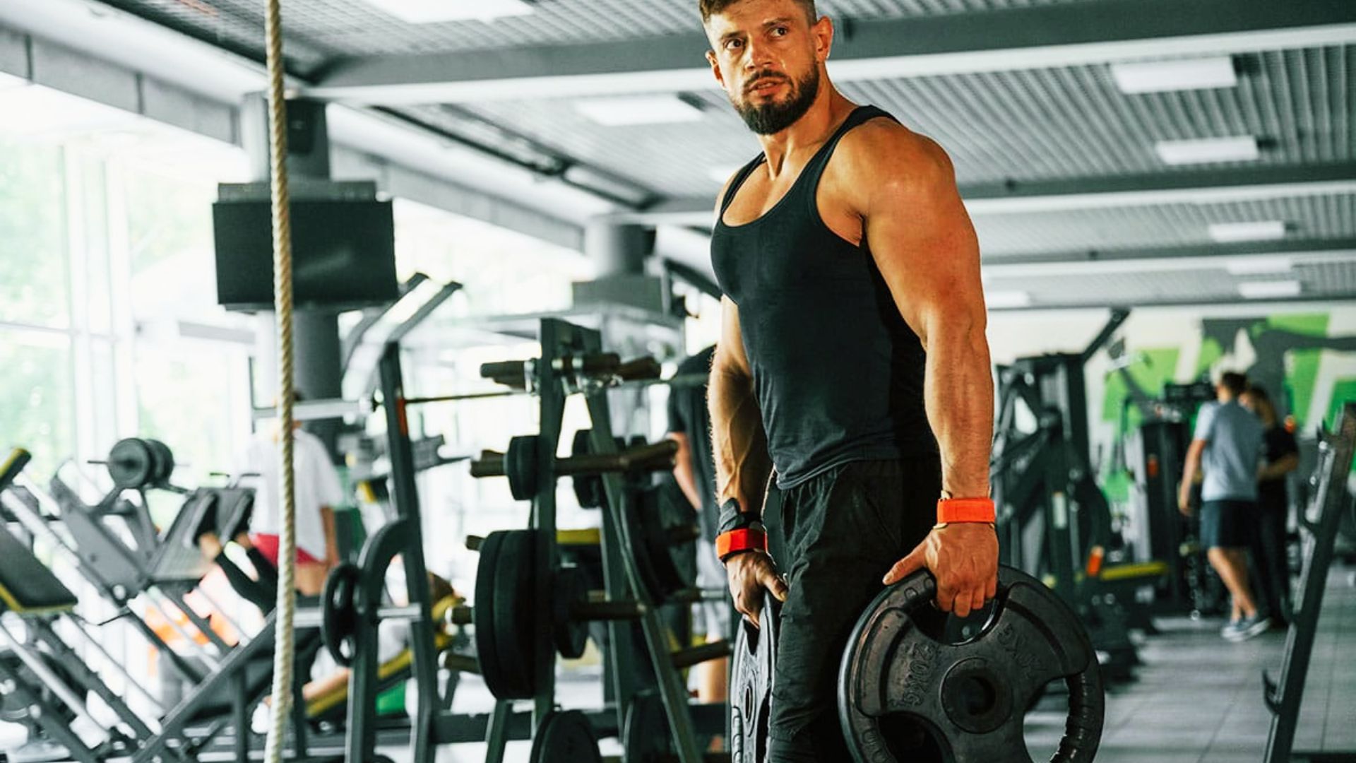 Muscular man in black tank top stands in the gym holding weight plates.