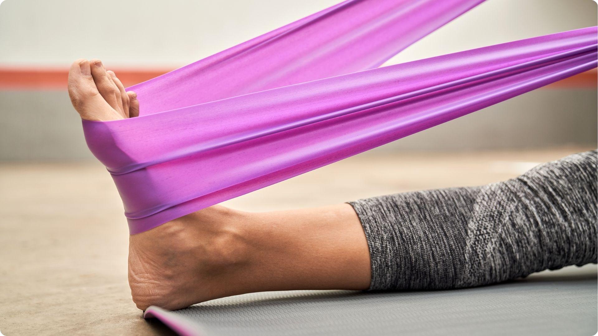 A person seated on a yoga mat pulls a purple resistance band wrapped around the toes to stretch the foot and calf.