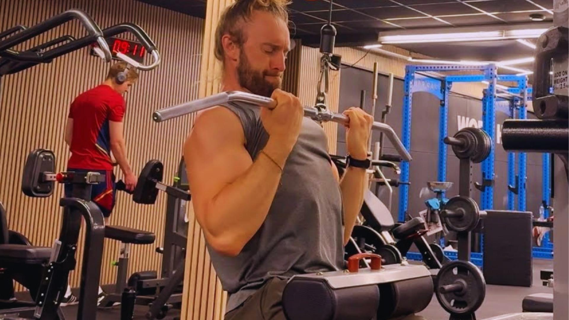 Bearded man in gray tank top performs supinated lat pulldown with underhand grip pulling bar to upper chest in modern gym.