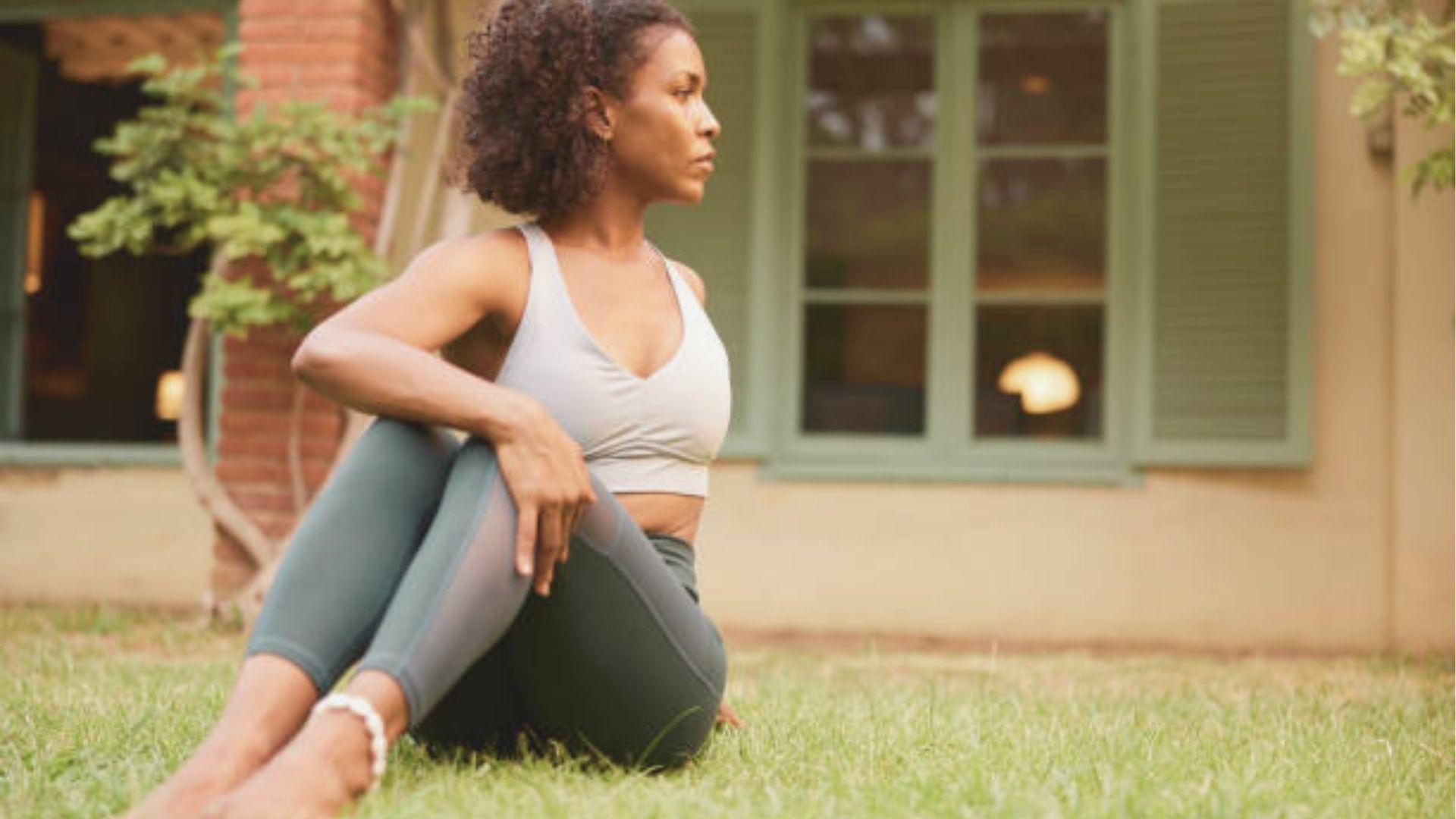 Woman in white sports bra and green leggings sits on grass in a seated spinal twist.