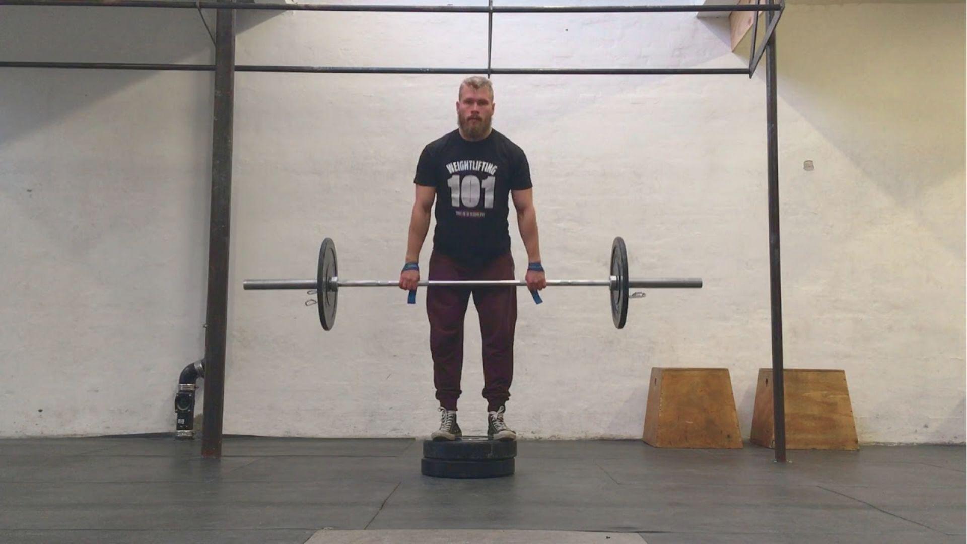 Bearded man in “Weightlifting 101” shirt standing on weight plates while holding a loaded barbell at lockout in a gym.