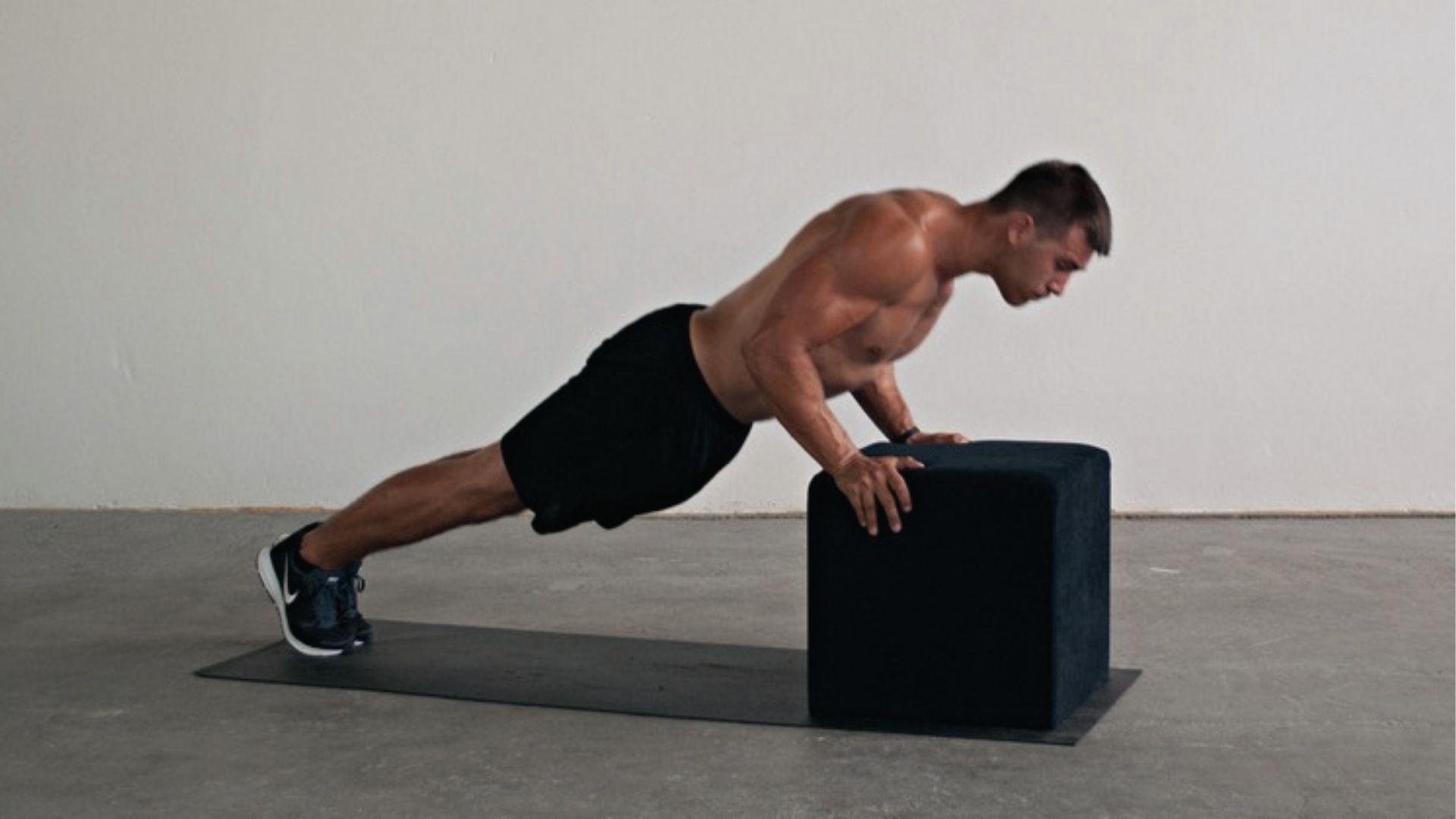 Shirtless man does incline push up using black box on yoga mat in minimal studio with white walls