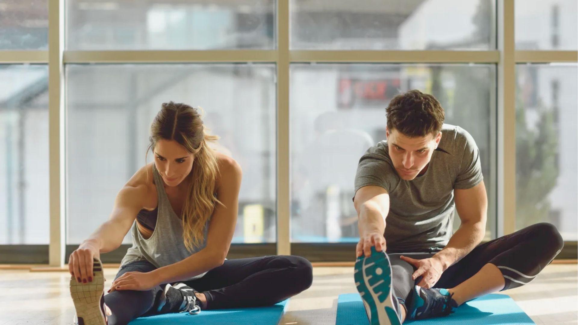 Man and woman sit on yoga mats in a bright studio, doing seated hamstring stretches by reaching for their feet.