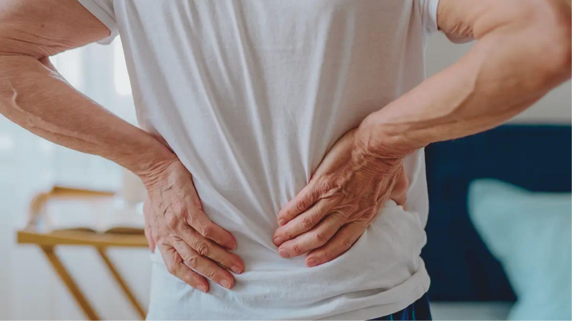 Older adult in a white t-shirt grips his lower back with both hands in a bedroom.