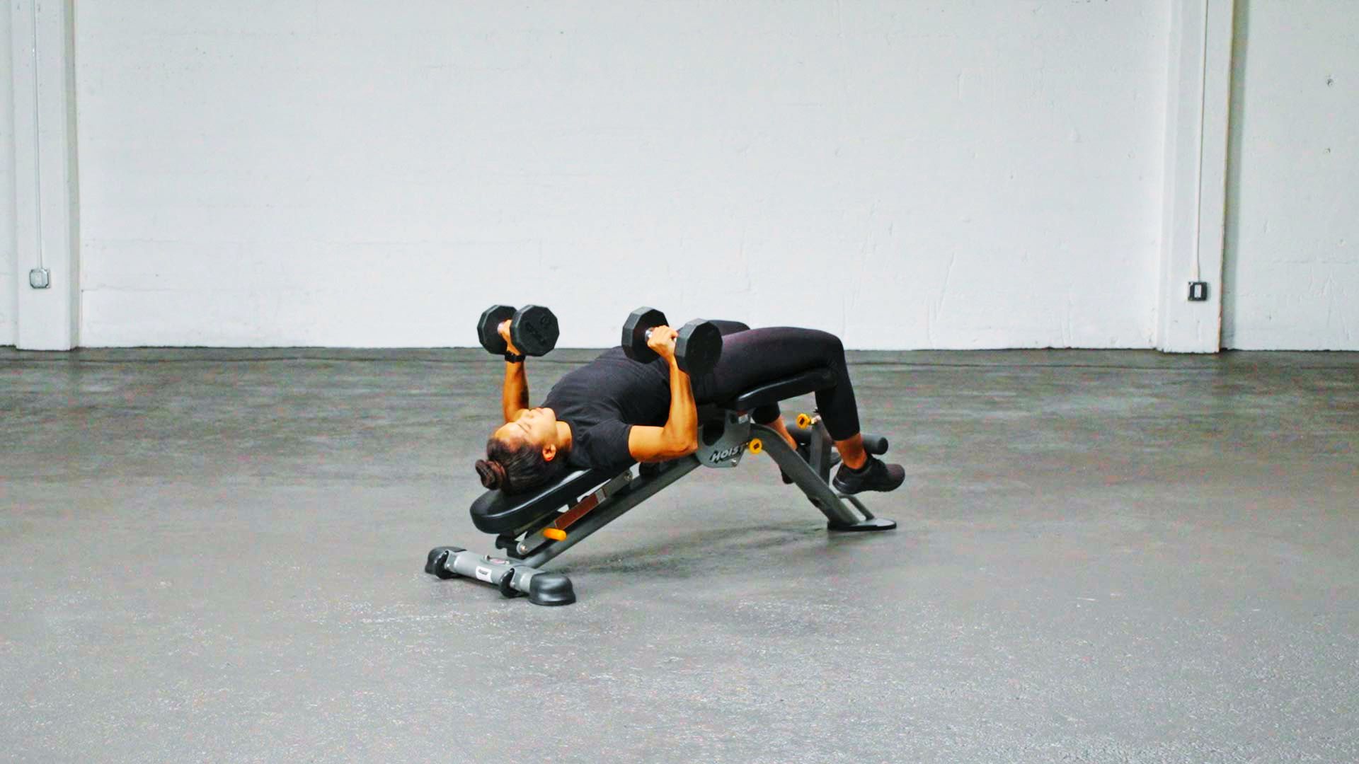A woman performs a decline dumbbell bench press on an angled bench with her head lower than hips.