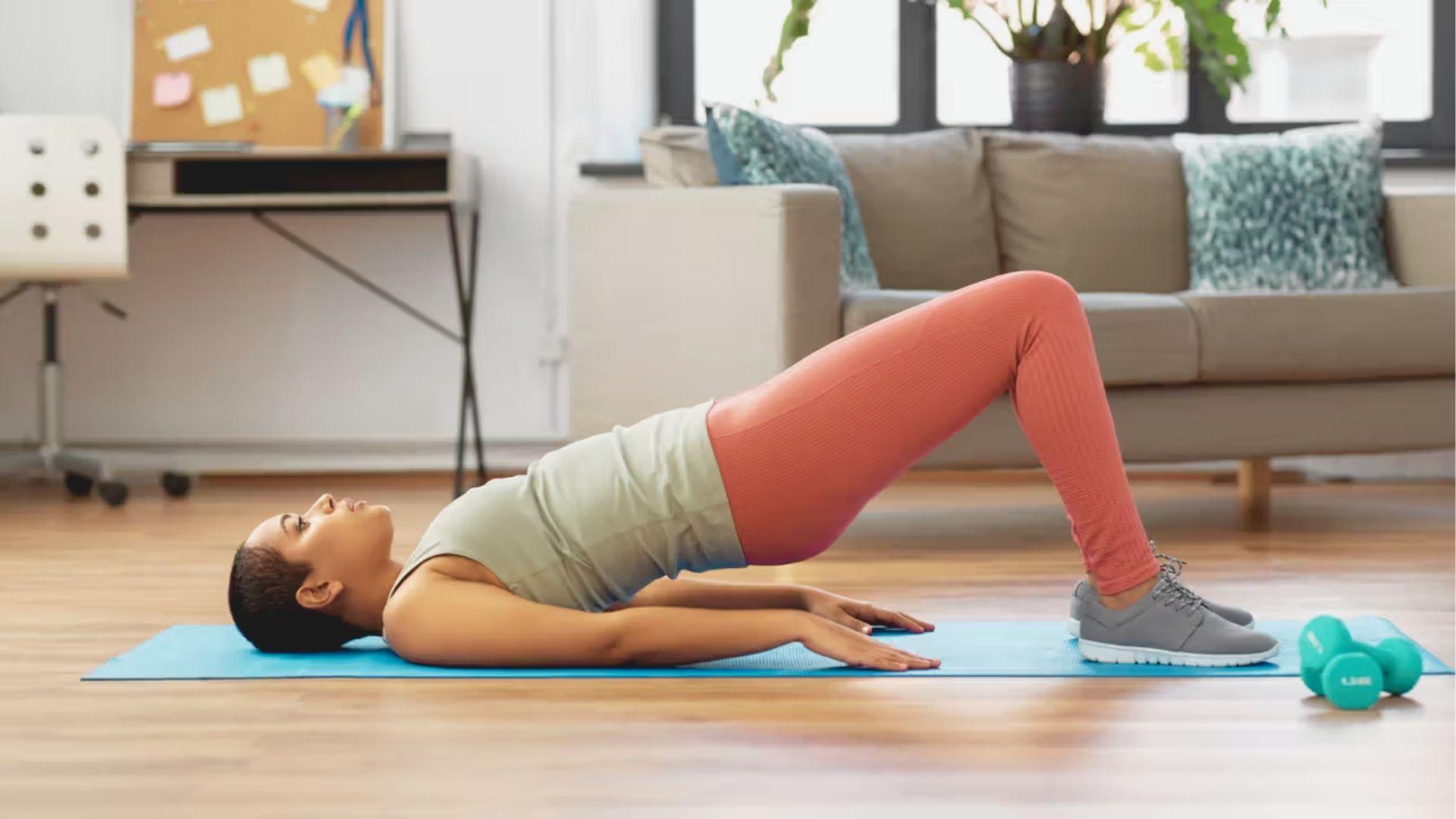 Woman performs a glute bridge exercise on a blue yoga mat at home with dumbbells nearby.