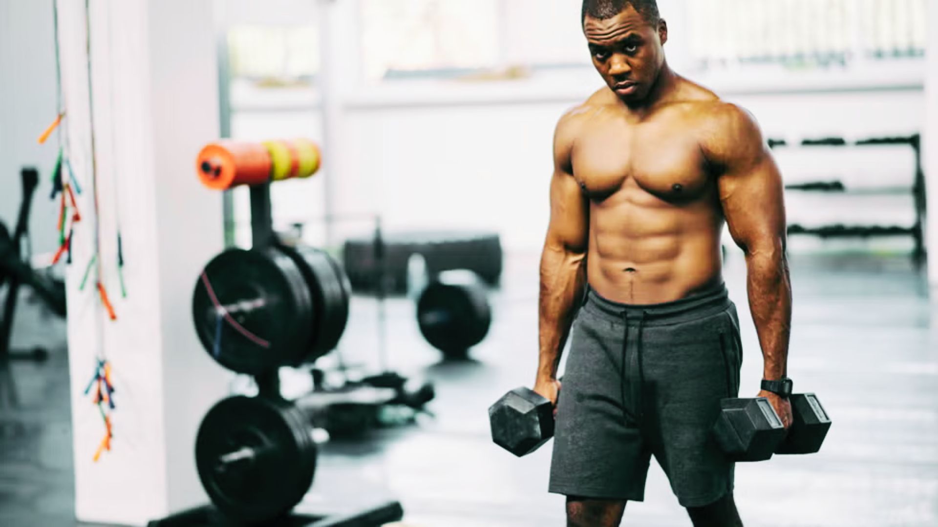 Focused, lean man with defined abs holding dumbbells mid-workout in a bright gym.