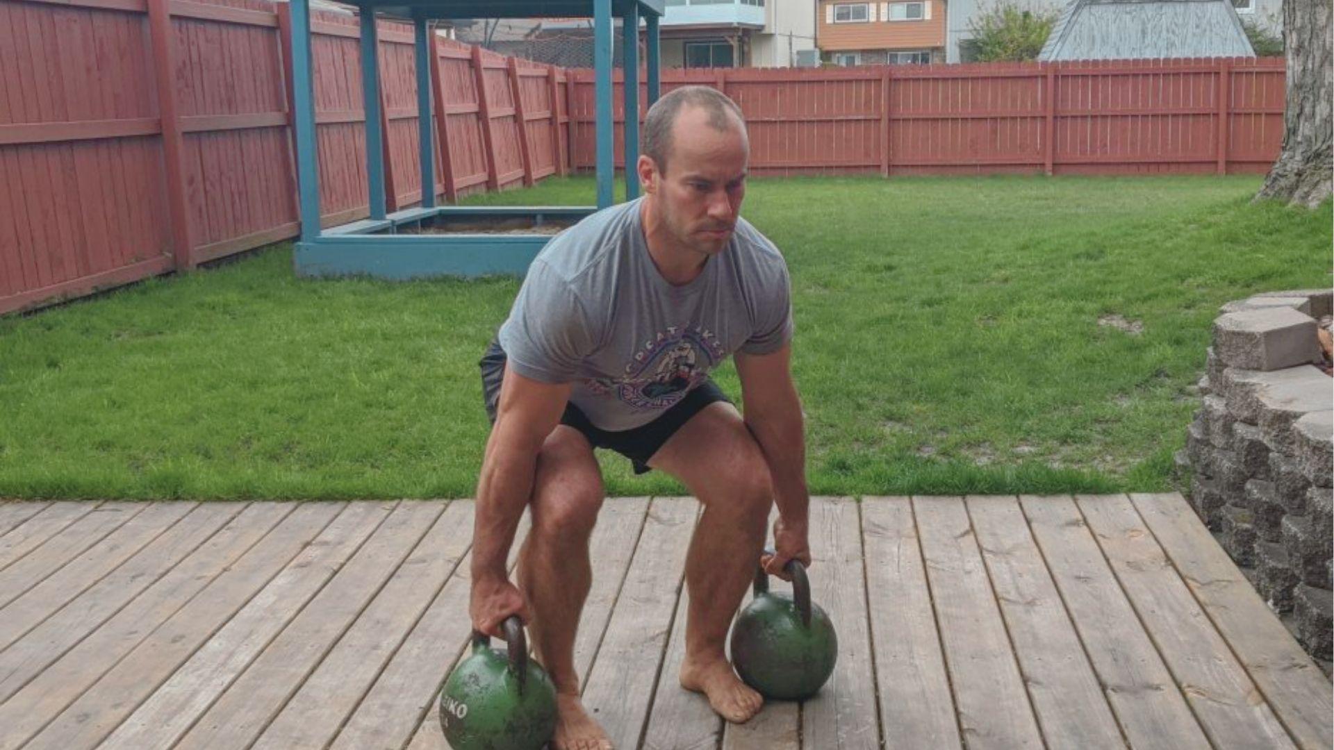 Man outdoors on a deck performing a dual kettlebell suitcase deadlift, barefoot in a squat stance.