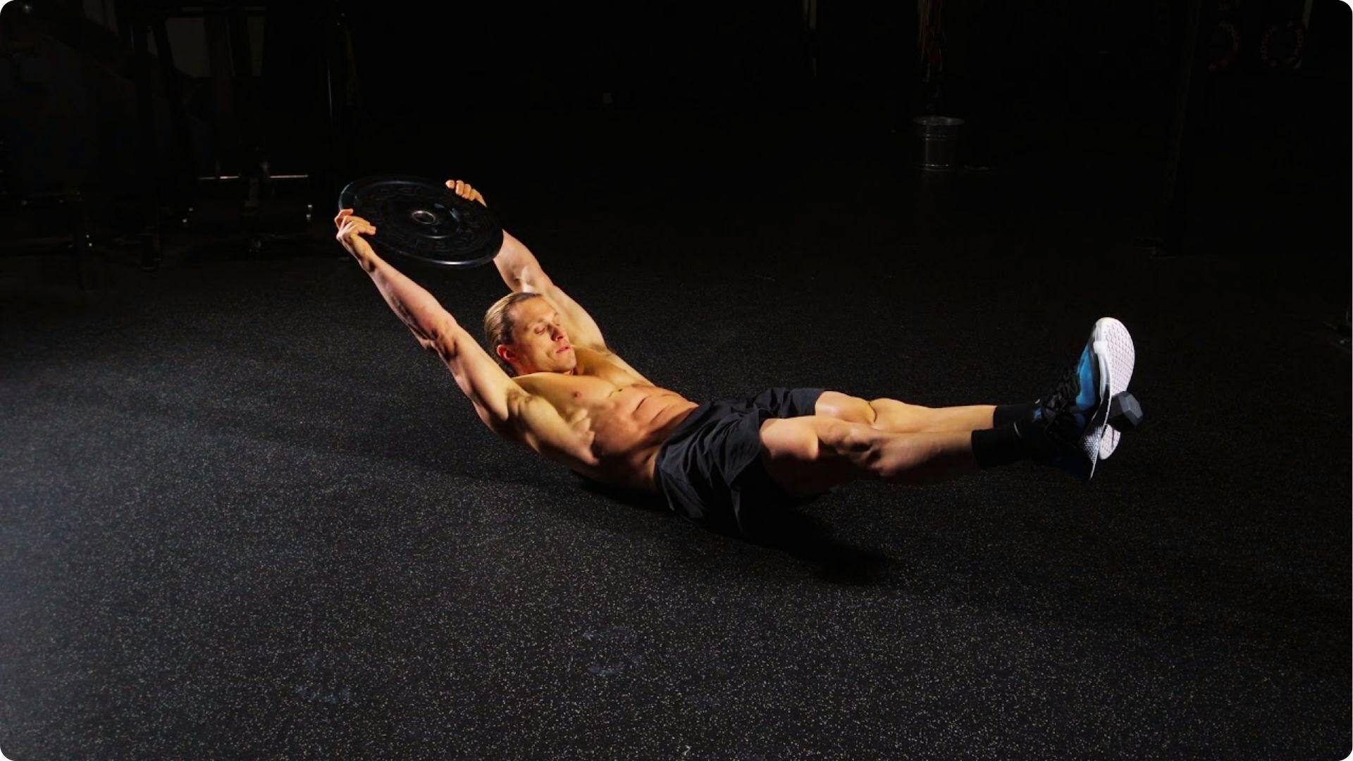A shirtless man performs a weighted hollow body hold on a dark gym floor.