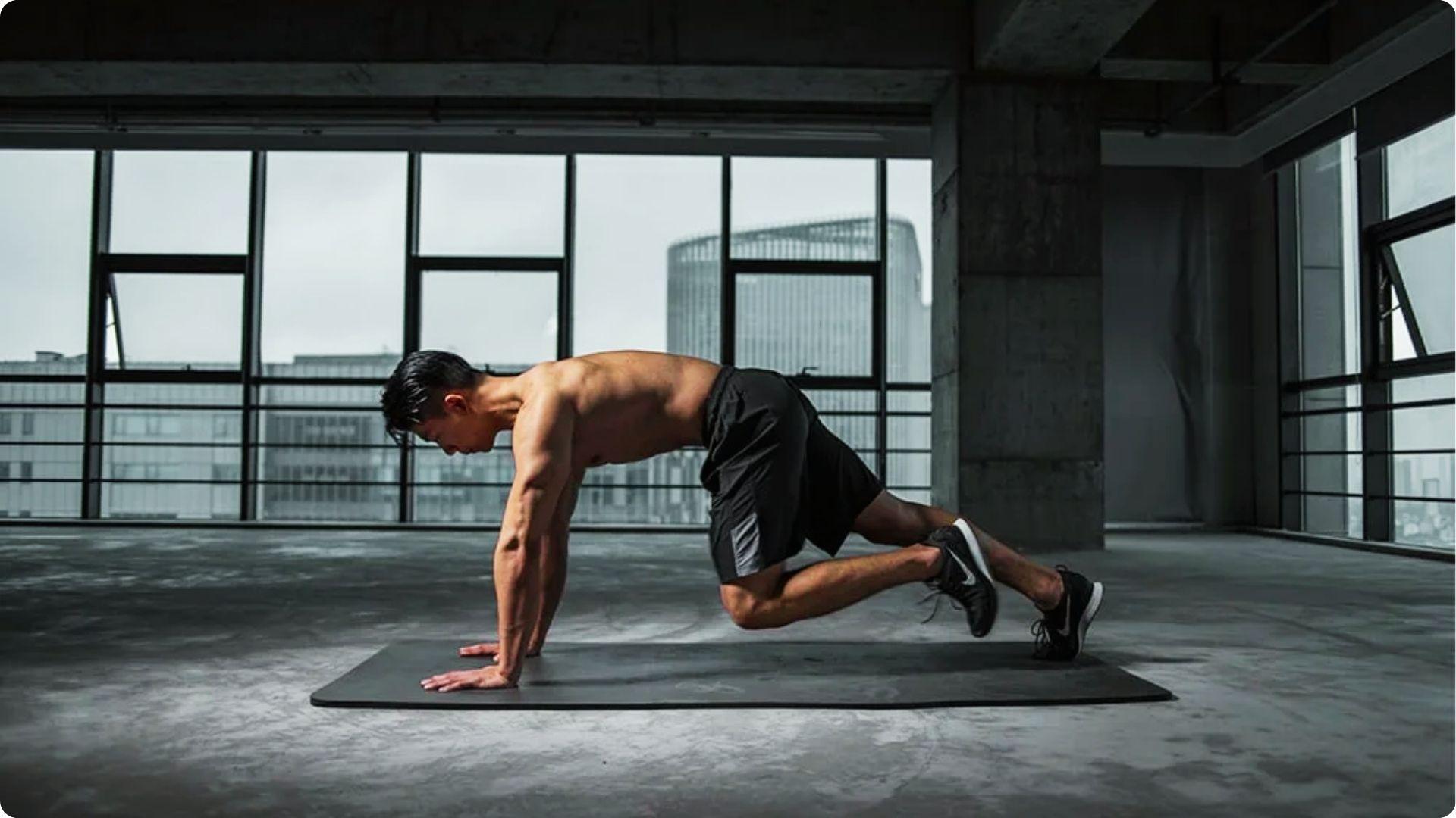 A shirtless man performs mountain climbers on a mat in an industrial space with large windows.