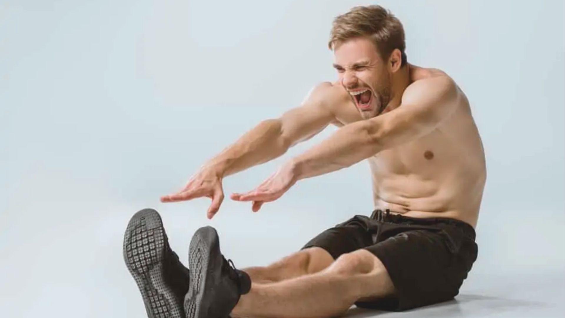 Shirtless man sits on the floor with legs extended, grimacing and reaching for his feet.