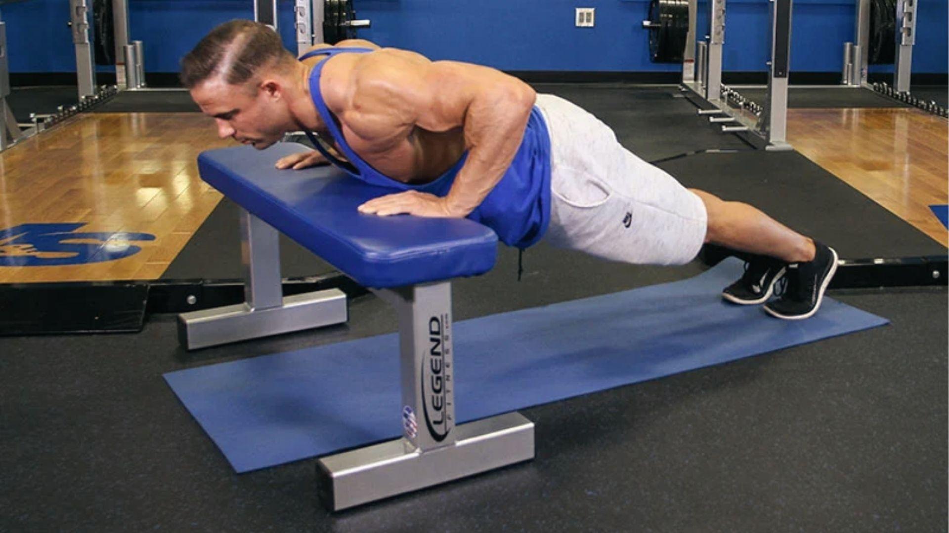 Muscular man in blue tank top does incline push up on blue bench over mat in modern gym