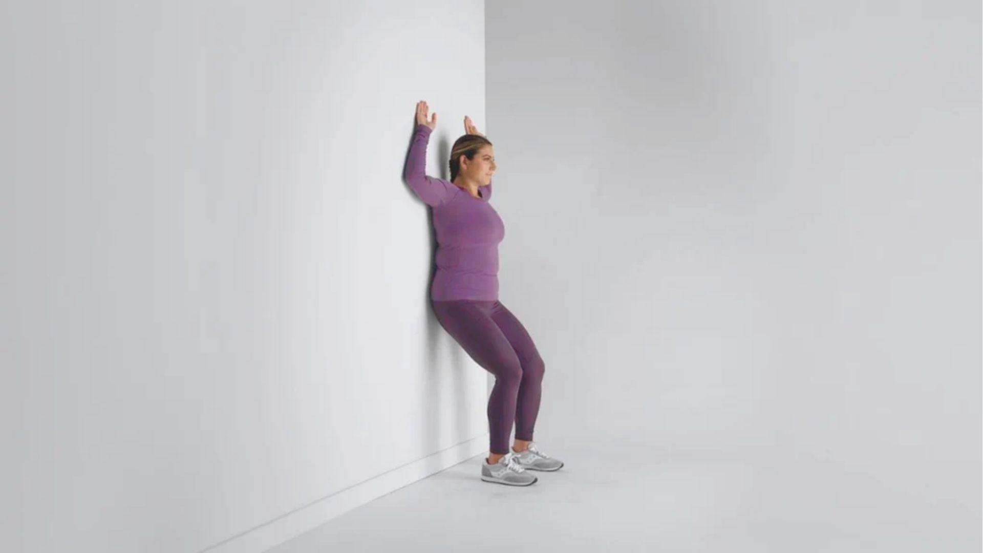 Woman in purple activewear performs a wall slide with arms raised against a white wall.