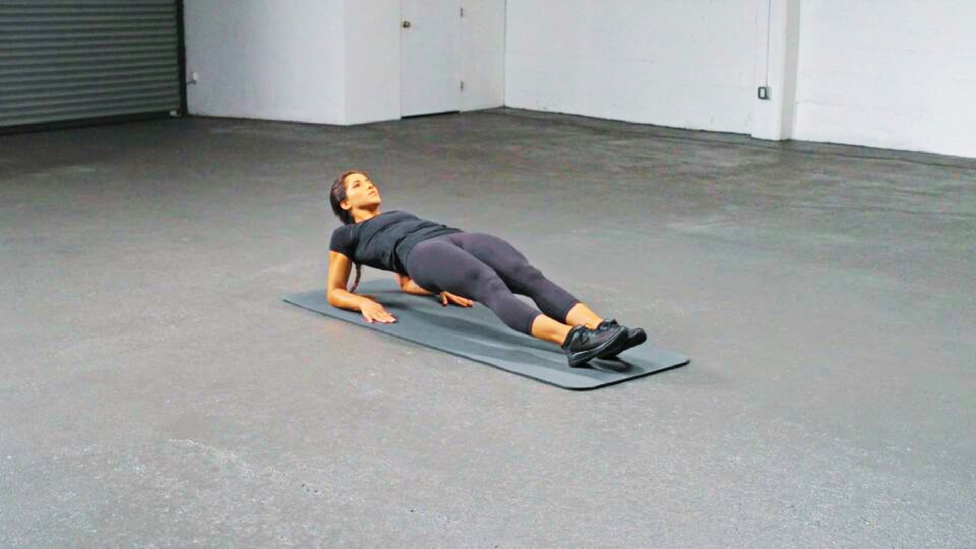 A woman in black workout wear holds a reverse plank on a yoga mat in an empty gym.