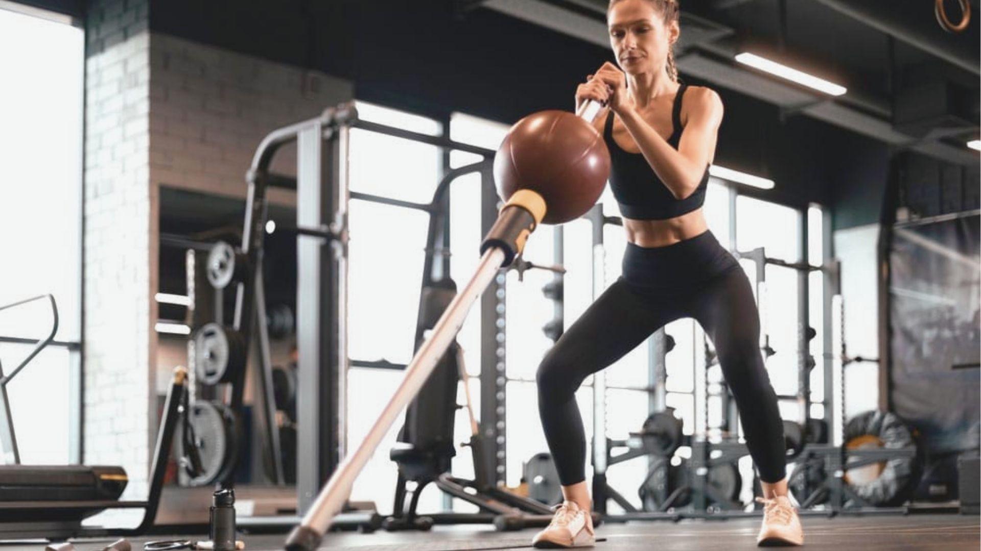 A woman in a black sports bra performs a wide-stance landmine squat using a barbell with a ball attachment.