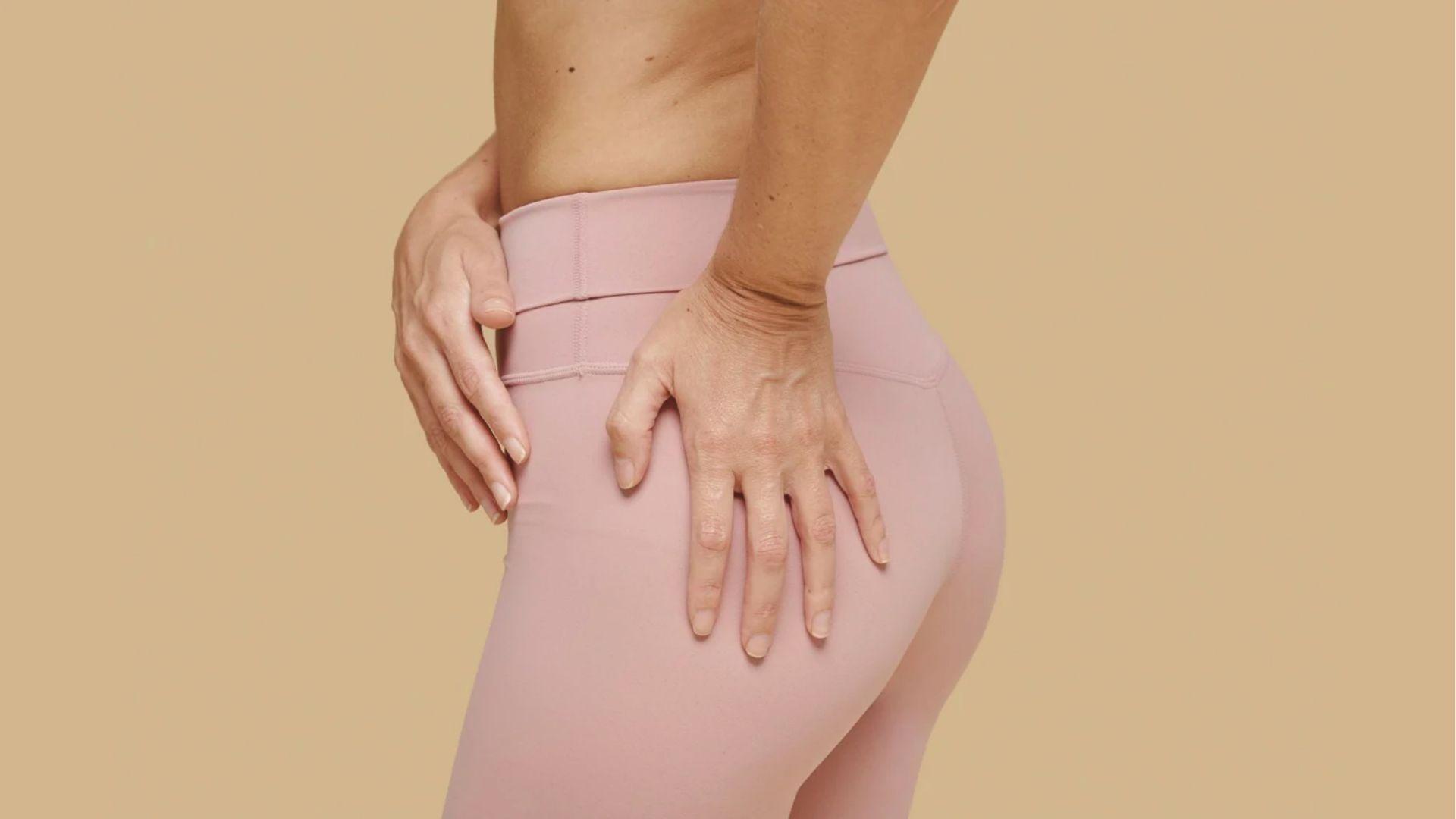 Woman in pink leggings placing hands on hips and glutes against a beige background.