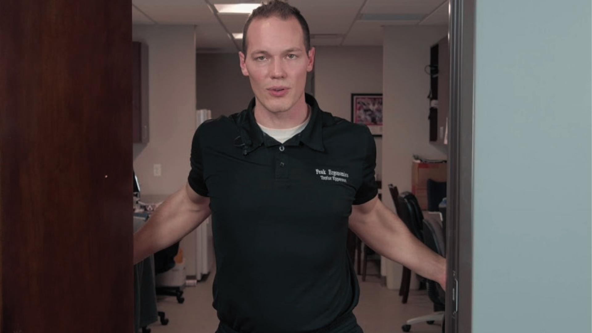 Man in black polo demonstrates doorway stretch for shoulder mobility, hands on doorframe during physical therapy.